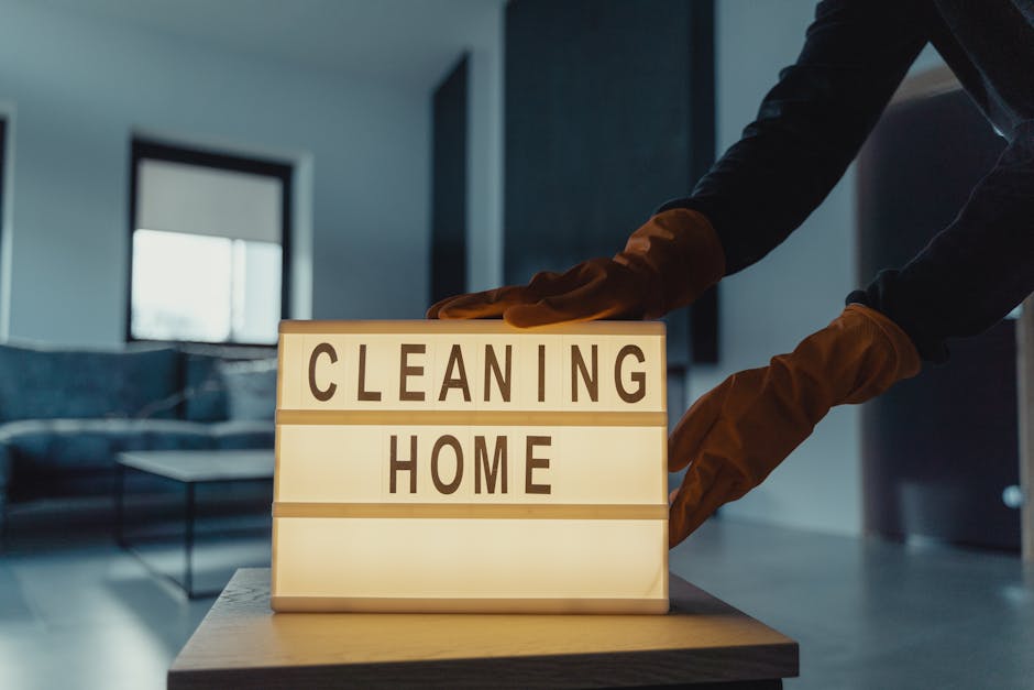 A person wearing brown cleaning gloves is adjusting a illuminated lightbox on a wooden table, which displays the words 'CLEANING HOME' in bold uppercase letters. The room has a modern interior with a
