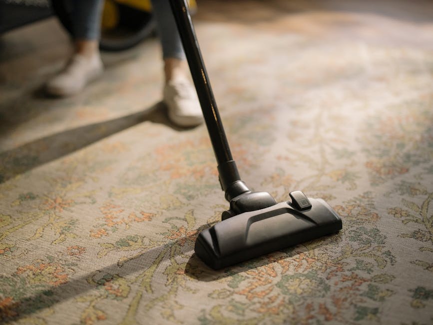 A yellow canister vacuum cleaner with a flexible hose and metal wand attachment rests on an ornate, patterned area rug in a living room. Nearby is a wooden sideboard with black metal legs, decorated w